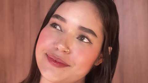 Media: Video of a young woman with light skin, dark brown hair, and a gold nose ring, smiling against a wooden background.