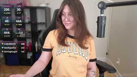 Video of a young woman with long brown hair, glasses, and a yellow T-shirt, seated in a gaming chair, streaming on Twitch. Background includes a black desk, monitor, and shelves with gaming accessories.
