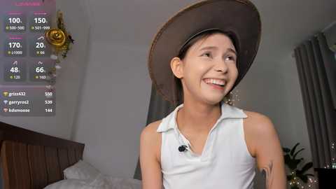 Video of a young woman with light skin, smiling in a white sleeveless polo shirt, wearing a large brown cowboy hat. Background includes a bed with white sheets, a plant, and a digital display showing game stats.