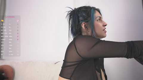 Video of a woman with blue-streaked hair in a sheer black top, facing right, against a white wall with a calendar.