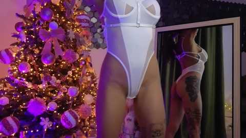 Video of a woman in white lingerie standing in front of a decorated Christmas tree, partially visible in a mirror, reflecting her body.