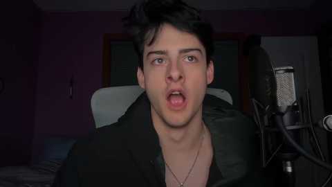 A video of a young man with short dark hair and a surprised expression, wearing a black shirt, in a dimly-lit bedroom with purple walls and a large silver microphone.