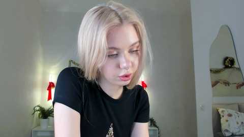 Video of a young woman with short, platinum blonde hair, wearing a black t-shirt, looking down, with red ribbons on her shoulders, in a cozy, softly lit room with a mirror and potted plants.