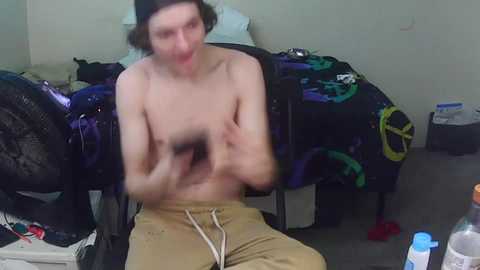 Video of a shirtless, fair-skinned, androgynous person with short, dark hair, wearing beige shorts, holding a black object. The background shows a messy bedroom with a bed, clothing, and a water bottle.