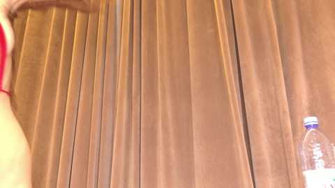 A video of a light-skinned person with a red bra strap partially visible, standing in front of a brown pleated curtain, with a clear plastic water bottle in the right foreground.