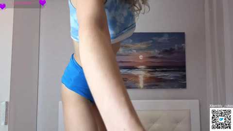Video of a woman in blue tie-dye sports bra and matching shorts, standing side profile. Background features a sunset painting and a white bed.