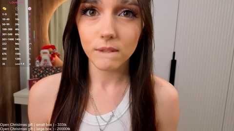 Video of a young woman with long dark hair, wearing a white tank top, and a silver necklace, standing indoors with a festive background.
