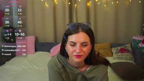 Media: Video of a young woman with long brown hair, wearing sunglasses and an olive-green hoodie, seated on a bed with a striped blanket. Background includes a cozy room with curtains, pillows, and fairy lights.