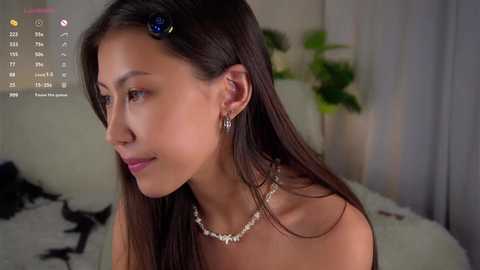 Video of an Asian woman with long black hair, blue hair clip, wearing a silver necklace and earrings, sitting in a softly lit bedroom, background blurred.
