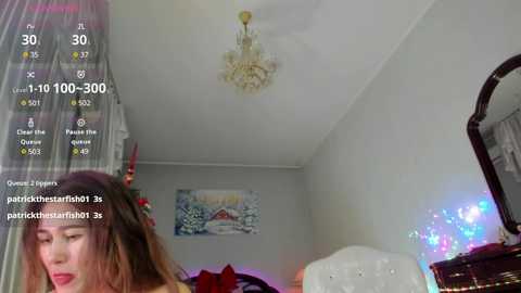 A video of a woman with long, straight brown hair, wearing a red dress, captured in a dimly lit bedroom with a chandelier, a dresser, and a bed with a red blanket.