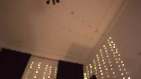 A dimly lit video shows a room with a chandelier, white ceiling, and a wall adorned with numerous small, glowing fairy lights, creating a cozy ambiance. Dark curtains partially obscure the view.