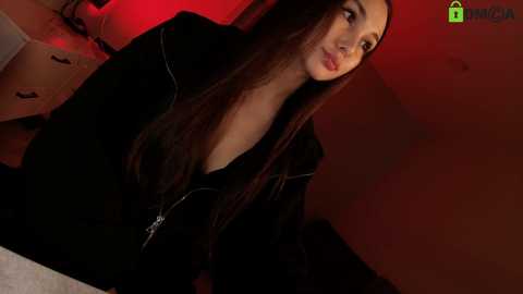 Media: A video of a young woman with long dark hair, wearing a black zip-up hoodie, sitting on a bed in a dimly lit room with red lighting.