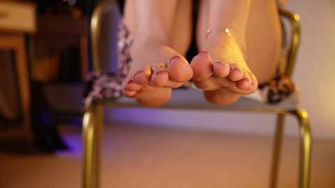 A video of a person's feet, toenails painted red, resting on a gold chair in a dimly lit room with a wooden table in the background.