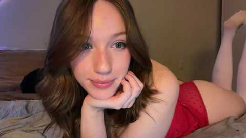 Video of a young, fair-skinned woman with long, wavy brown hair, wearing red lace lingerie, lying on a bed. She has a soft smile and rests her chin on her hands.