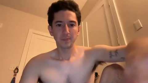 Video of a shirtless, fair-skinned man with short dark hair, wearing a neutral expression, standing in a beige room with a white door and light switches visible.