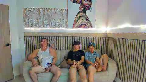 Video of a young family sitting on a light beige sofa with wooden paneling. The father, wearing a white tank top and shorts, sits on the left, while the mother in a black shirt and the son in a blue shirt sit in the center. A large framed painting of a black woman with braids is on the wall behind them.