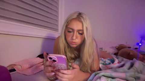 A video of a blonde, fair-skinned young woman lying on a bed, texting on a pink phone. The room is dimly lit with purple light, and stuffed animals are scattered around.