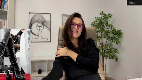 Video of a woman with long brown hair, wearing red glasses and a black outfit, sitting in an office chair in front of a computer screen, surrounded by minimalist decor, including a plant and artwork on the wall.