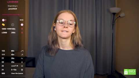 Video of a young, light-skinned woman with glasses and long, straight, light brown hair, wearing a gray sweater. She stands against a gray curtain backdrop. The image is from a live stream, with chat and viewer count displayed.