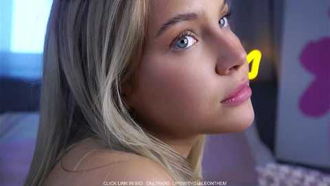 Video of a close-up of a young woman with light skin, straight blonde hair, blue eyes, and a neutral expression, set in a dimly lit room with blurred abstract art and a bed in the background.
