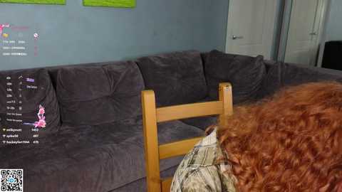 Video of a woman with voluminous, curly red hair, sitting on a dark gray sectional sofa, facing a wooden chair, in a simple, modern living room.