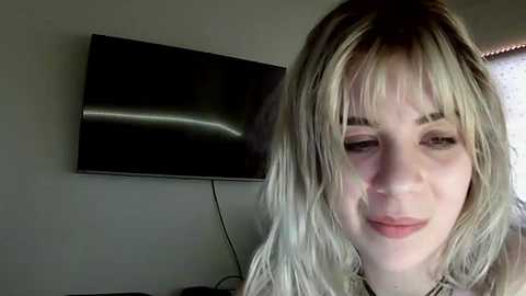 A video of a young woman with long, wavy blonde hair and fair skin, wearing a light-colored top, smiling softly. A large black TV is mounted on a wall behind her.