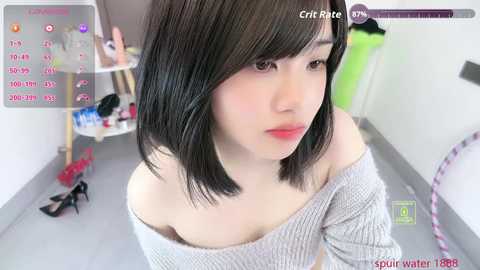 A video of a young East Asian woman with short black hair, fair skin, and red lipstick, wearing a gray off-shoulder knit top. She stands in a tidy, brightly lit room with a white table and a pink chair in the background.