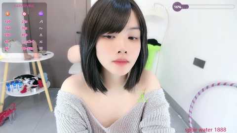 A young Asian woman with short black hair, light skin, and a slender physique, wearing a gray off-shoulder sweater, sits in a modern, minimalistic room with a white chair, laundry basket, and a colorful toy on the floor.