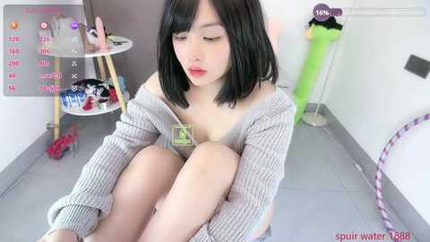 A video of an Asian woman with short black hair, wearing a grey sweater, sitting on the floor, looking down, with a small green card in her hand.