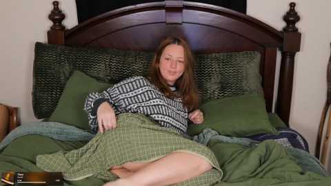 Video of a Caucasian woman with straight brown hair, lying in bed, wearing a green and black patterned sweater, covered with a green plaid blanket, on a green bedspread.