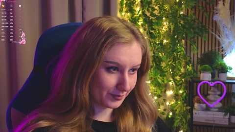 A video of a smiling young woman with long, wavy blonde hair, wearing a black top, seated in a dark blue gaming chair. A green wall with fairy lights and a purple heart-shaped neon light is in the background.