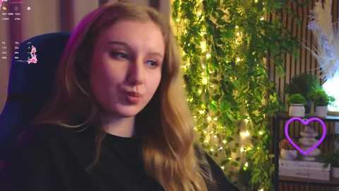 A video of a young woman with long, wavy blonde hair, fair skin, and a black top, making a kissing gesture, in a cozy, dimly lit room with greenery and fairy lights.