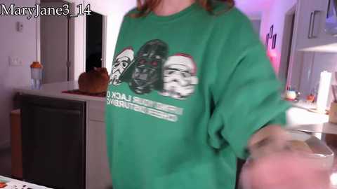 A video of a person wearing a green Star Wars-themed sweatshirt featuring Darth Vader, Stormtrooper, and Boba Fett. The background shows a modern kitchen with a countertop, orange cup, and a candle.