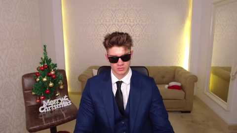 Video of a man in a navy suit, black tie, and sunglasses, seated in a modern, dimly-lit living room with a Christmas tree and \"Merry Christmas\" sign, beige couch, and white wallpaper.