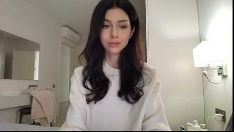 Video of a fair-skinned woman with long, wavy dark brown hair, wearing a white sweater, sitting in a modern bathroom with white walls, mirror, and a wall-mounted light.