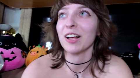 Video of a young woman with shoulder-length brown hair, light skin, and blue eyes, wearing a black choker, in a room filled with Halloween-themed decorations, including a pumpkin and plush toys.