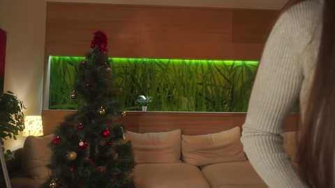 Video of a cozy living room with a Christmas tree adorned with red and gold ornaments, a beige couch, and a large aquarium with green plants.