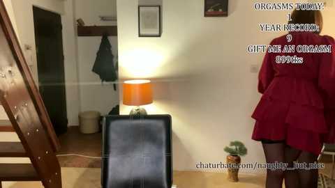 Video of a woman in a red dress, standing near a lamp and a potted plant in a cozy living room. Text overlays mention \"Orgasms Today\" and \"Gifts for Organisms.\
