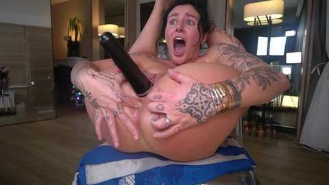 Video of a tattooed man with a shocked expression, holding a black dildo, sitting on a blue chair in a modern, brightly lit room.