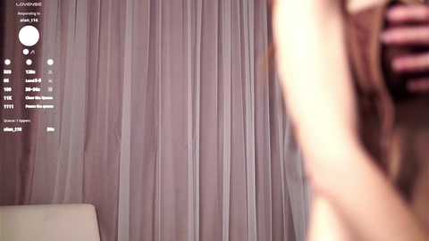 A blurred video showing a partially visible person with long hair in front of sheer, light purple curtains. The background includes a white couch and a digital camera display with settings.