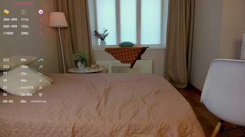 A video of a cozy, modern bedroom with beige walls, a queen-sized bed with a tan quilt, and a white chair in the foreground. The room features a window with sheer curtains and a wooden floor.