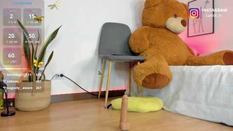 Video of a plush teddy bear on a gray chair in a cozy, minimalist bedroom with a wooden floor, a potted plant, and a white bedsheet.