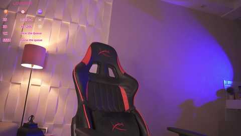 A video of a modern gaming chair with red accents, positioned in a dimly lit room with a beige wall, a table lamp, and a purple-blue light source.