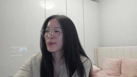 Video of an Asian woman with long black hair, wearing glasses, a beige cardigan, and a pink shirt, smiling in a modern, minimalist bedroom with white walls and a cream-colored headboard.