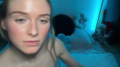 Video of a young, fair-skinned woman with blonde hair, looking contemplative, in a dimly lit room with blue lighting. Behind her, a large stuffed animal and a small Christmas tree.