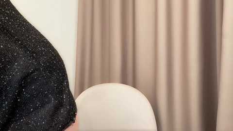 Video of a person in a black, glittery top and beige curtain, seated on a white chair with a smooth, curved back.