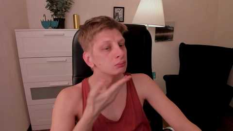 Video of a young, fair-skinned man with short, light brown hair, wearing a red tank top, sitting in a black chair, scratching his neck, in a beige room with white drawers, a lamp, and a plant.