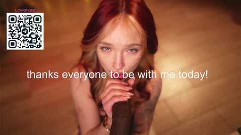 Video of a nude woman with long, red hair, kneeling on a wooden floor, holding a dildo, and saying, \"thanks everyone to be with me today!\