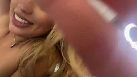 Video of a smiling woman with light skin and blonde hair, wearing a gold necklace, taken from a low angle.