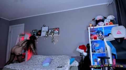 A video of a nude woman with long dark hair and slender physique, bending over a bed, in a dimly lit, cluttered bedroom with various toys, decorations, and red lighting.